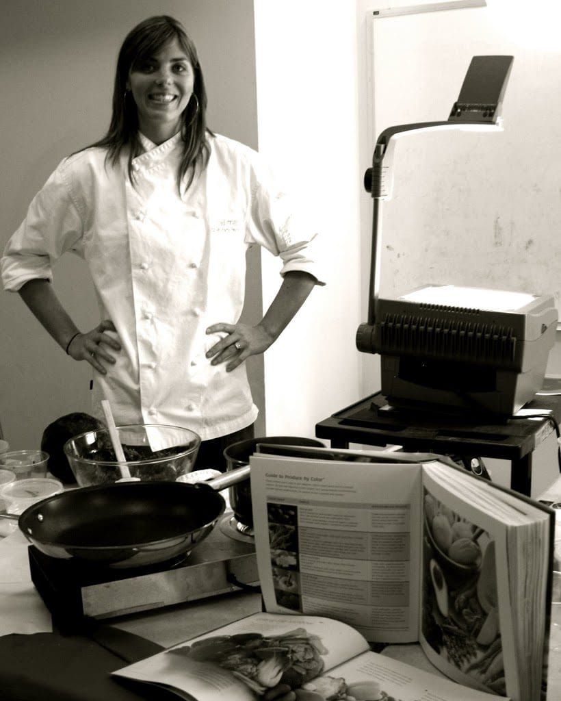 A woman in a chef's uniform standing in front of a pan.